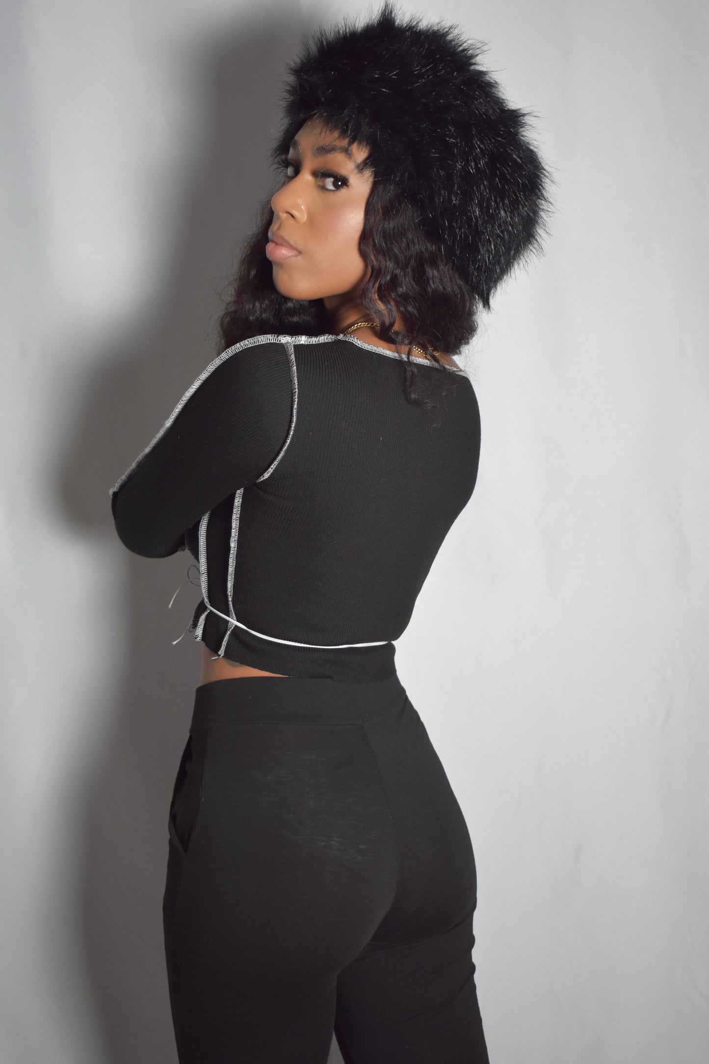 Back view of solid long-sleeve crop top with no lace-up detail, bold stitching around shirt, black faux fur headband, black pants, y2k streetwear style