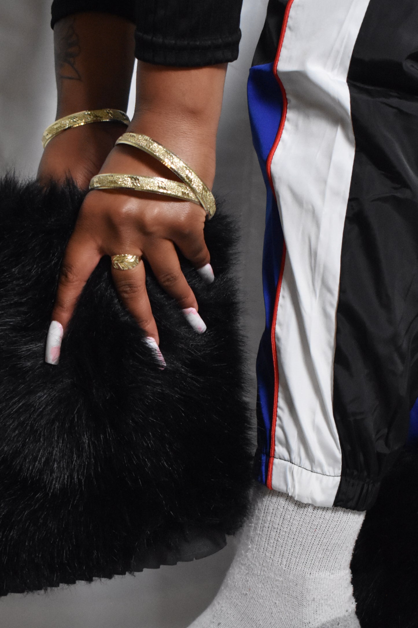 Hand with gold bracelets and ring on a black fur boot. black color block track pants with white stripe and yellow-blue panels, black faux fur boots, nostalgic y2k retro streetwear style