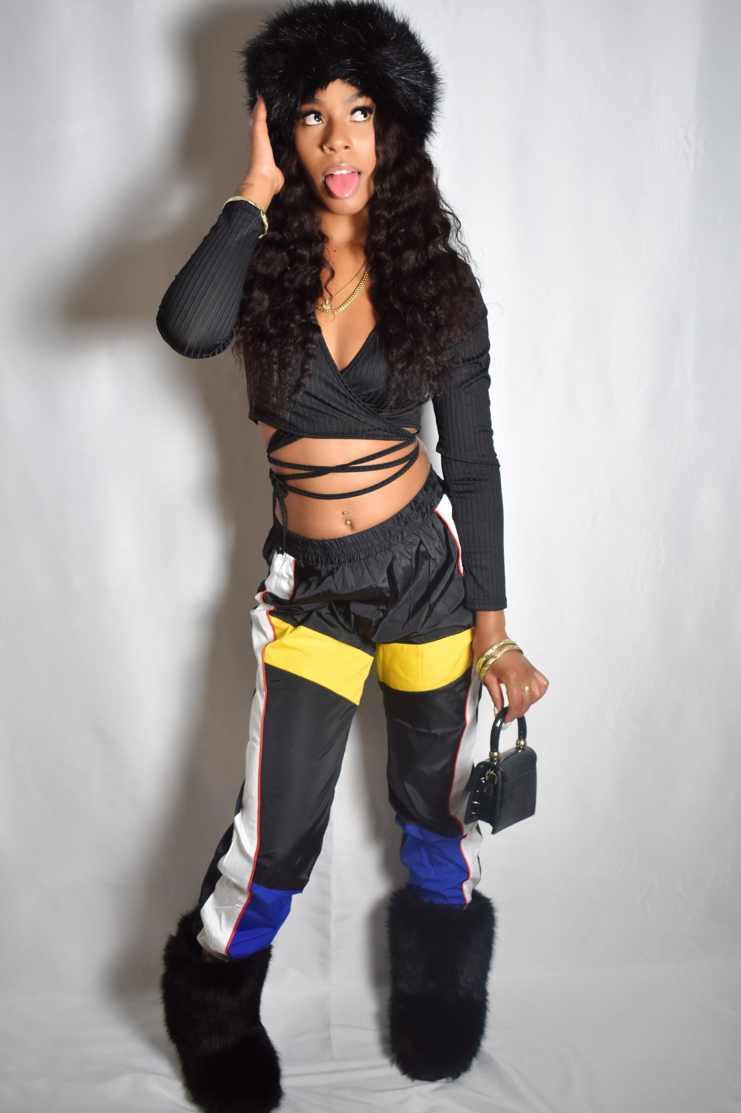Woman wearing a stylish outfit with a fur headband and black outfit on a plain background. black color block track pants with white stripe and yellow-blue panels, long sleeve wrap up customizable crop top, jumbo faux fur headband, black mini leather purse, black faux fur boots, nostalgic y2k retro streetwear style