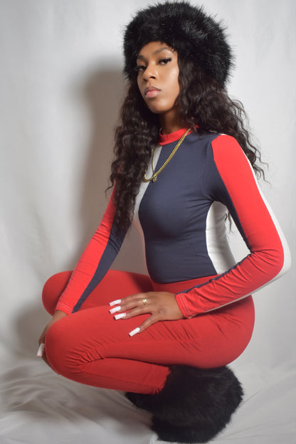 Woman wearing a red and navy outfit with a fur headband on a plain background. Arctic Rebel color block bodysuit in red, white, and blue with slim fit. Red stacked pants. Black jumbo faux fur headband and boot accents.