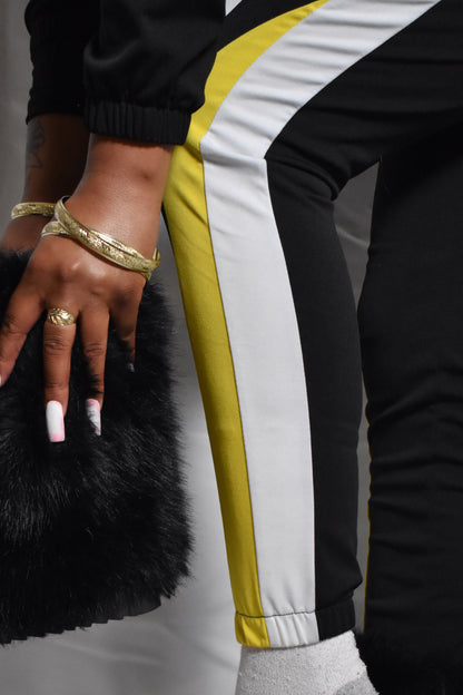 Person wearing a black tracksuit with yellow and white stripes, holding a black fur boot black retro y2k tracksuit color block with yellow and white accents, brown faux fur boots, brown jumbo faux fur headband, two piece outfit set