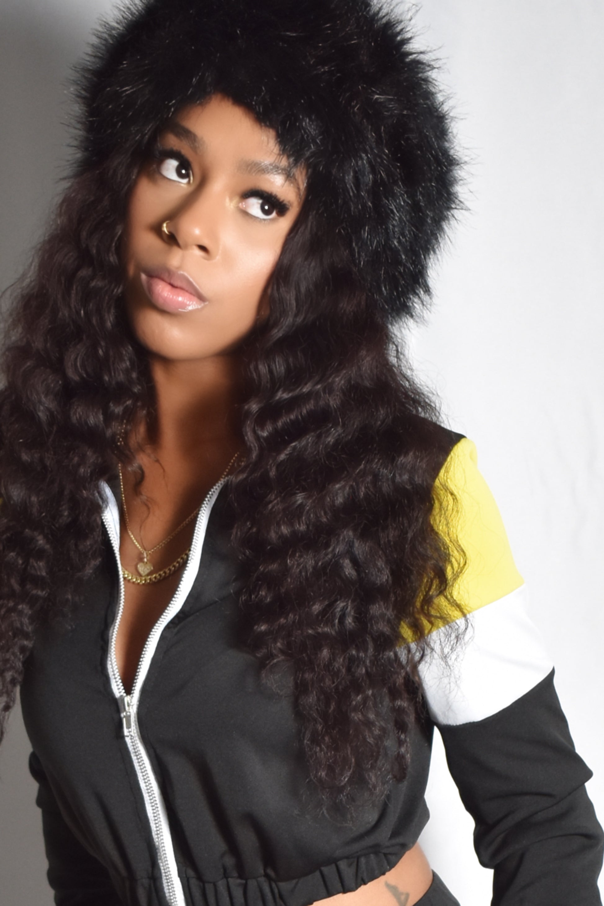 Woman wearing a black jacket with a fur-lined headband against a white background. black retro y2k tracksuit color block with yellow and white accents, brown faux fur boots, brown jumbo faux fur headband, two piece outfit set