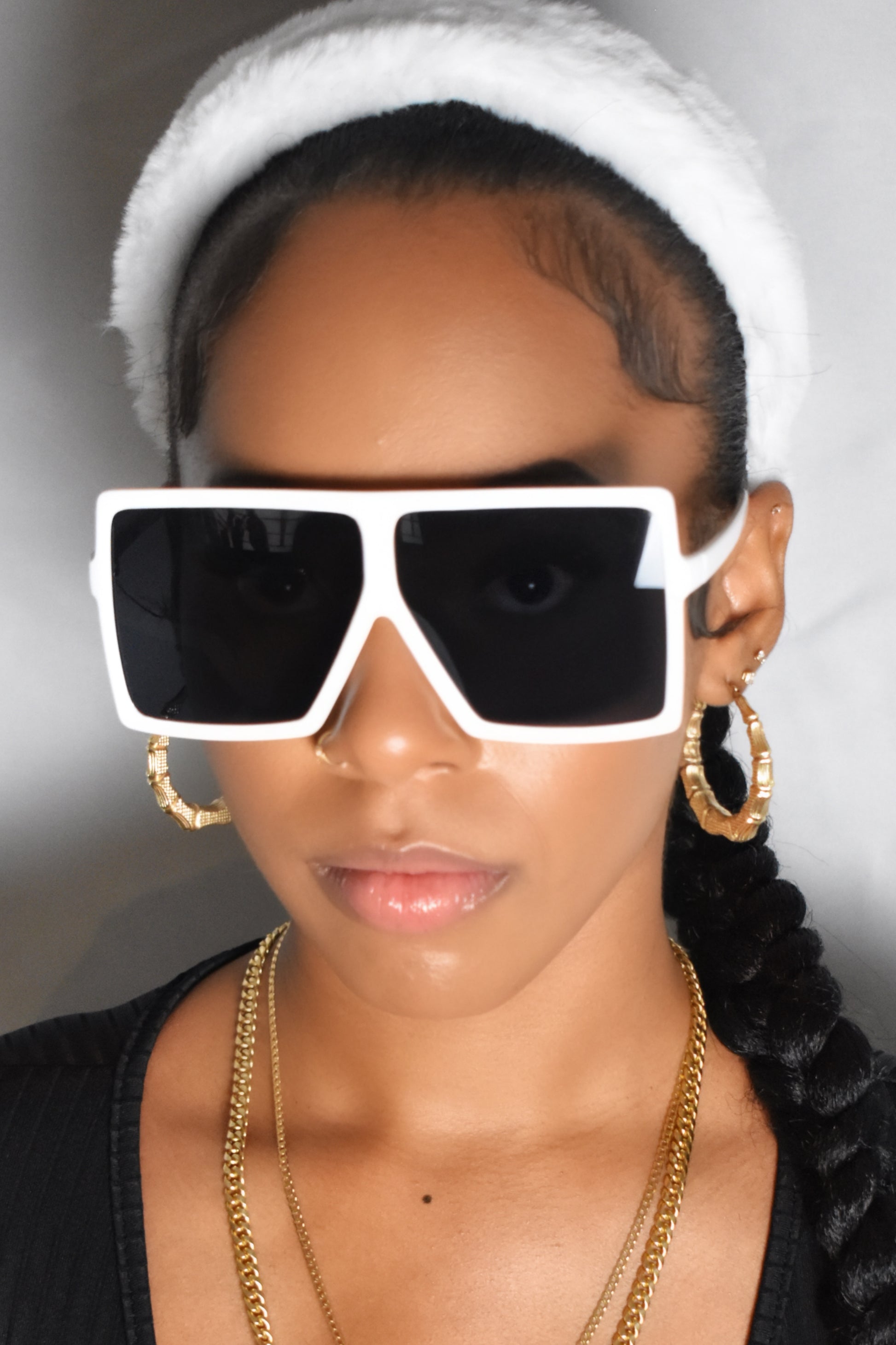 Person wearing large white sunglasses with black lenses, a white headband, and gold jewelry. Oversized white frame sunglasses with semi-dark tinted lenses. Close-up of white frame sunglasses with tinted lenses.