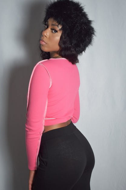 Back view of solid pink long-sleeve crop top with no lace-up detail, bold stitching around shirt, black faux fur headband, black pants, y2k streetwear style