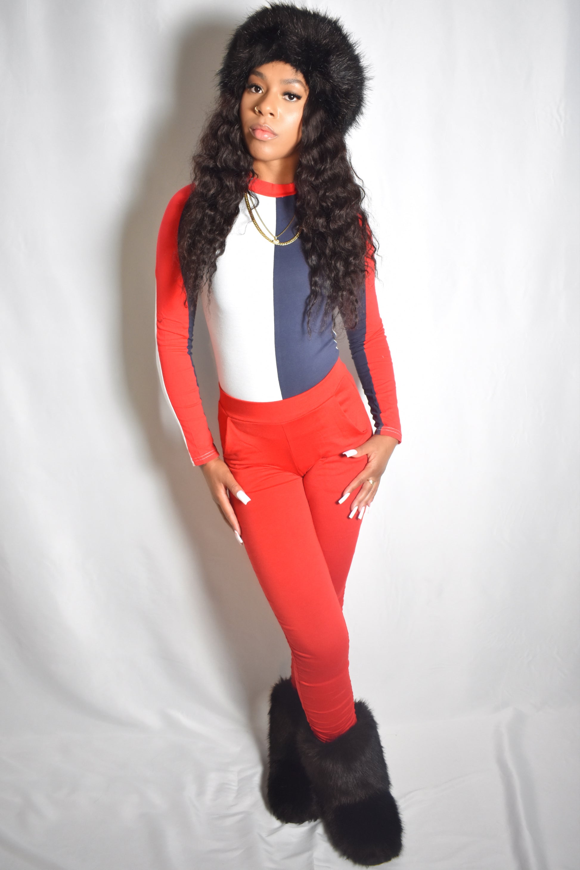 Person wearing a red and blue tracksuit with a fur-lined hood on a white background. Arctic Rebel color block bodysuit in red, white, and blue with slim fit. Red stacked pants. Black jumbo faux fur headband and boot accents.