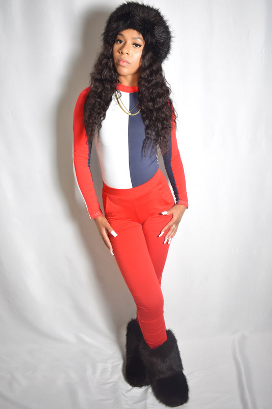 Person wearing a red and blue tracksuit with a fur-lined hood on a white background. Arctic Rebel color block bodysuit in red, white, and blue with slim fit. Red stacked pants. Black jumbo faux fur headband and boot accents.