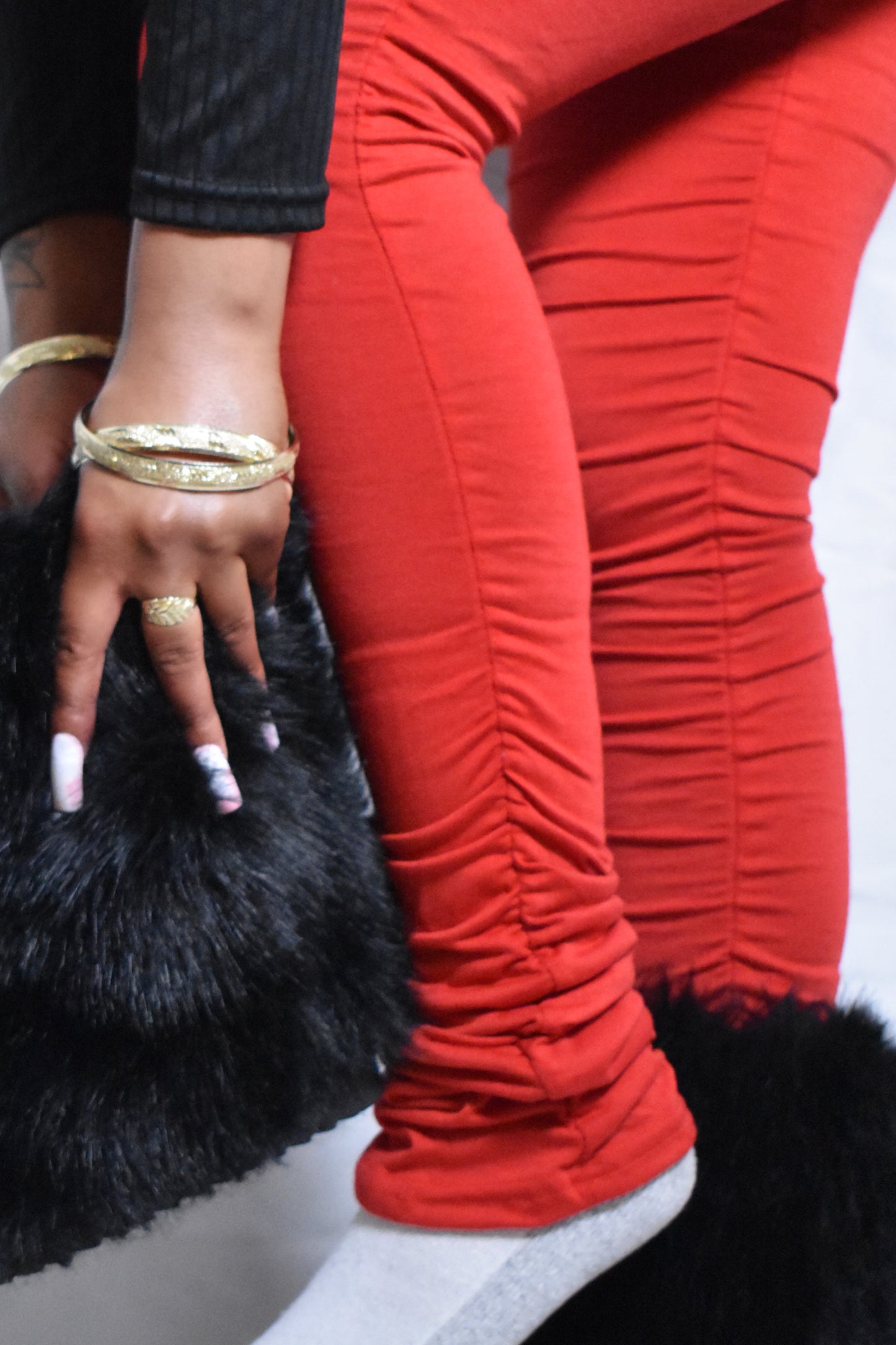 Person wearing red pants with gold jewelry. Model wearing red stacked pants with fitted silhouette. Black faux fur boots. Stacked pants hem.