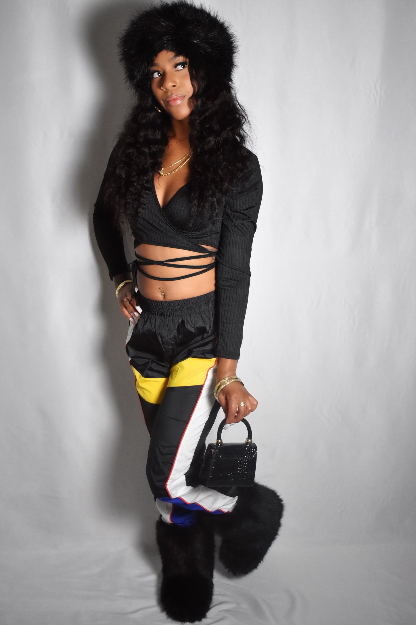Woman wearing a black outfit with fur details and holding a black handbag on a gray background. black outfit with colorful retro y2k color block pants and fur accents. Tie up wrap black crop top. Black fur faux boots. Black jumbo faux fur headband luxury. black crocodile-patterned handbag with gold accents. Essentials Only mini faux leather handbag in black, compact and chic design. textured faux leather and structured silhouette. sleek faux leather finish