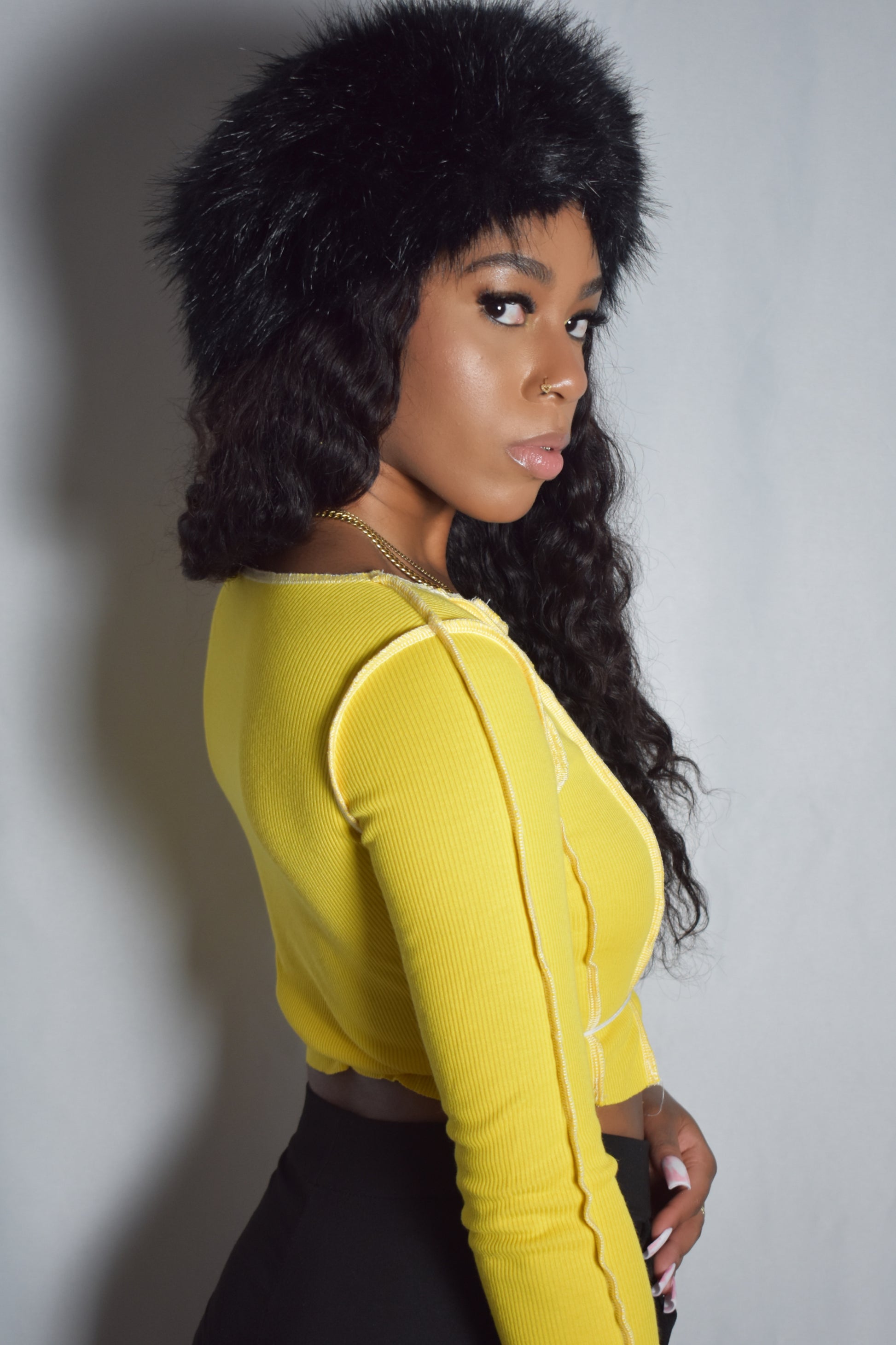 Back view of yellow solid long-sleeve crop top with no lace-up detail, bold stitching around shirt, black faux fur headband, black pants, y2k streetwear style